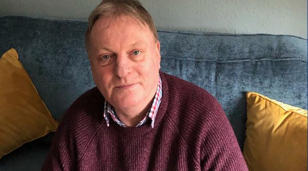 Paul Monaghan sits on a grey sofa with yellow cushions and looks at the camera. He is wearing a burgundy jumper and a checked shirt underneath.