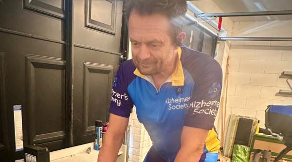 A man in Alzheimer's Society cycling shorts and t shirt on an excerise bike in a garage