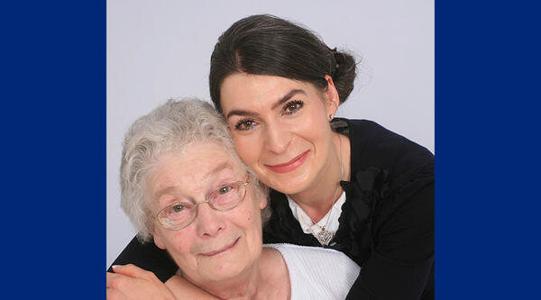 Tricia has dark brown hair, tied back, she's wearing a black jacket and she's hugging her mum who has short white hair, glasses and is wearing a white blouse