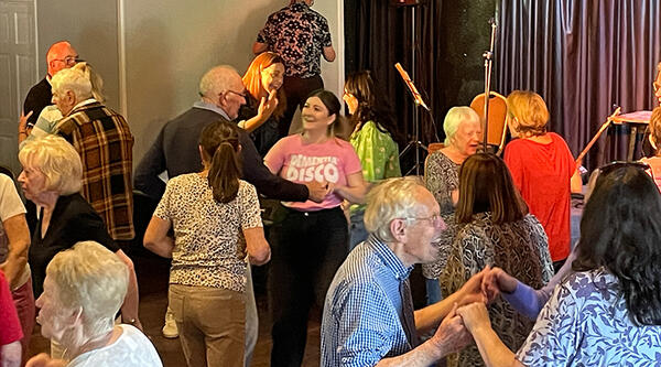Dancefloor filled with around 15 people dancing, including person wearing pink Dementia Disco t-shirt
