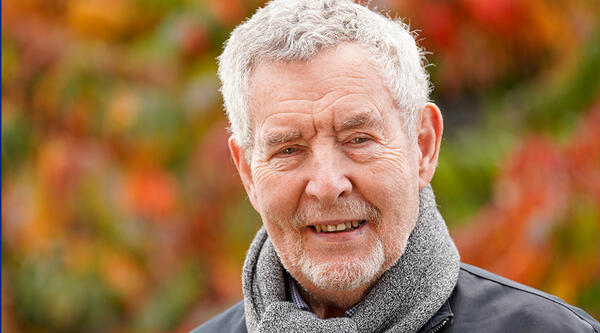 Ian Johns in coat and scarf, standing with autumnal trees behind him