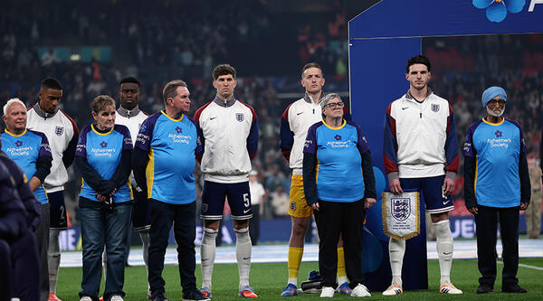 Susan wearing a blue Alzheimer's Society t-shirt, alongside other mascots and England football players on Wembley Stadium's pitch