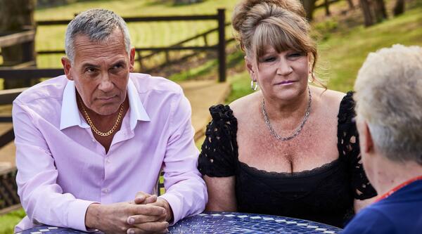 A man in a pink shirt with grey hair and a woman with in a black top with a blonde updo listening to a man with grey hair