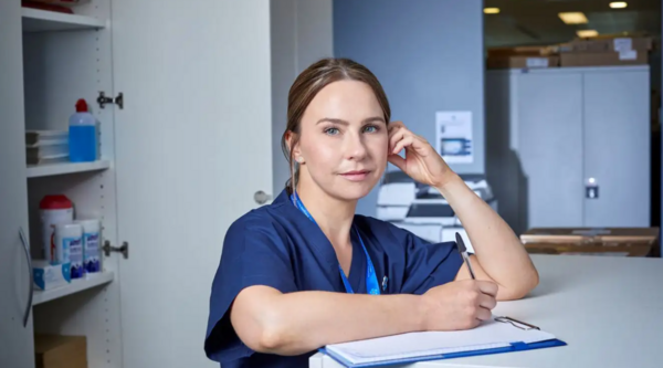 A nurse leans on a hospital counter with a clipboard
