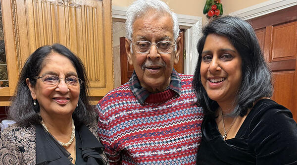 Nupur and her mum Protima, who are both wearing black clothes, are stood either side of her dad Bulu. He is wearing a red stripy jumper. They are stood in a kitchen. 
