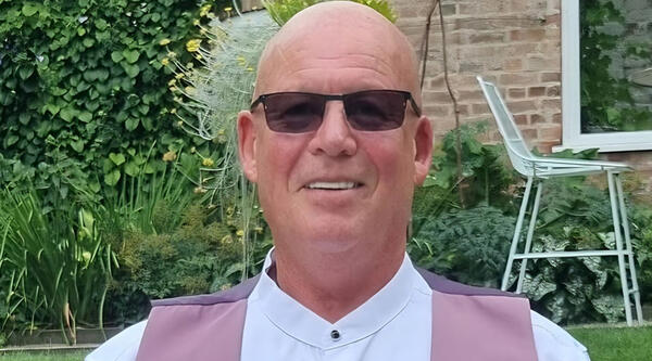 John is sat in a garden, wearing sunglasses, a white shirt and pink waistcoat.