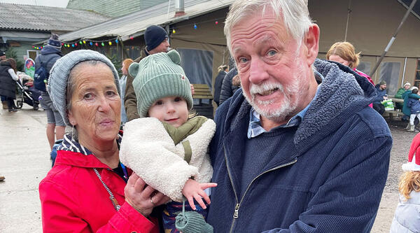 Ellen and her husband Malcolm. Ellen is wearing a red coat and grey hat. Malcolm is wearing a blue coat and holding a toddler.