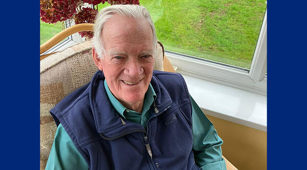 Philip is sat in an armchair, wearing a green shirt and blue gilet.
