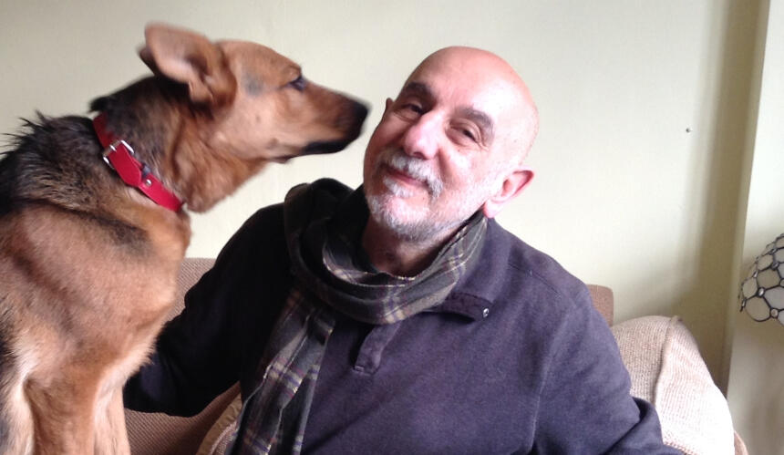 Armen sits on a sofa with a large brown dog holding its nose close to Armen's cheek, who is smiling
