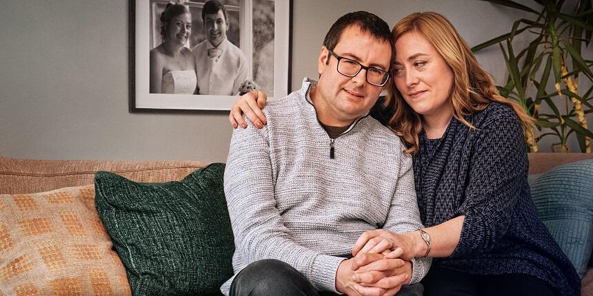 Mark and Caroline sit together on a sofa while holding hands