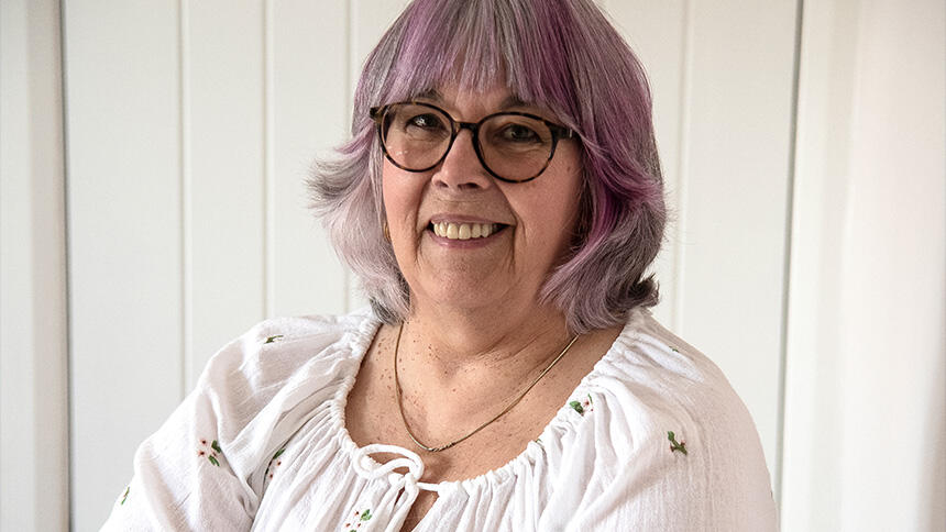 Gill Taylor is wearing glasses and a white blouse ad has purple hair. She stands in her home, cross armed, holding a paintbrush in her right hand, looking at the camera