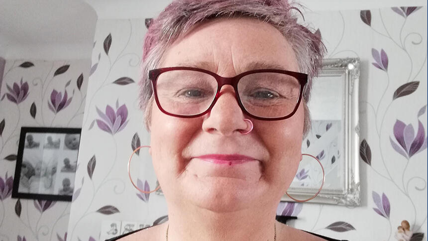 A head and shoulders shot of Michelle Nelson-Greensmith in her living room. She is smiling at the camera with pink hair and is wearing glasses.