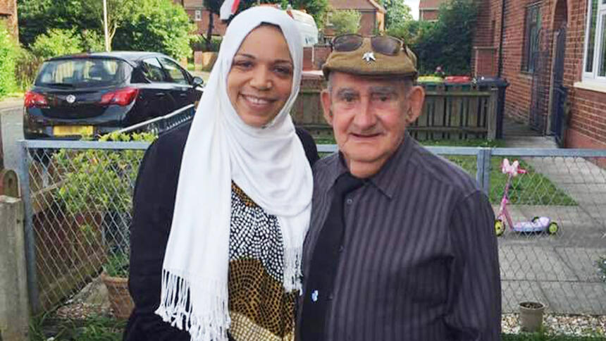 Jaymain and Bob smiling while stood outside a house, she is wearing a white headscarf, he is wearing a blue shirt and tie, and a flatcap
