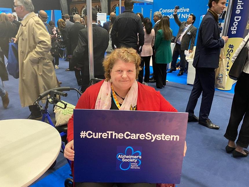 Julie at the conservative party conference holding a sign that says 'Cure the care system'
