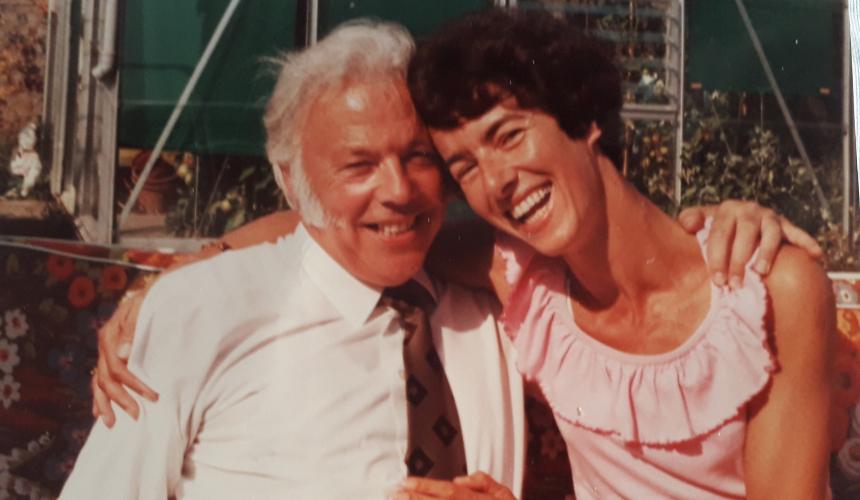 A grainy coloured photo of Doug wearing a white shirt and tie besides his wife Sylvia who is laughing with her hand on his chest