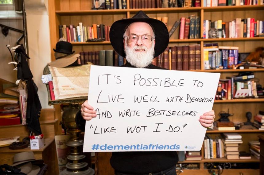 Terry Pratchett holding a Dementia Friends sign that reads 'It's possible to live well with dementia and write bestsellers "Like wot I do"!'