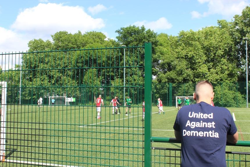 A person facing towards a football pitch where a game is happening, wearing an Alzheimer's Society branded t shirt
