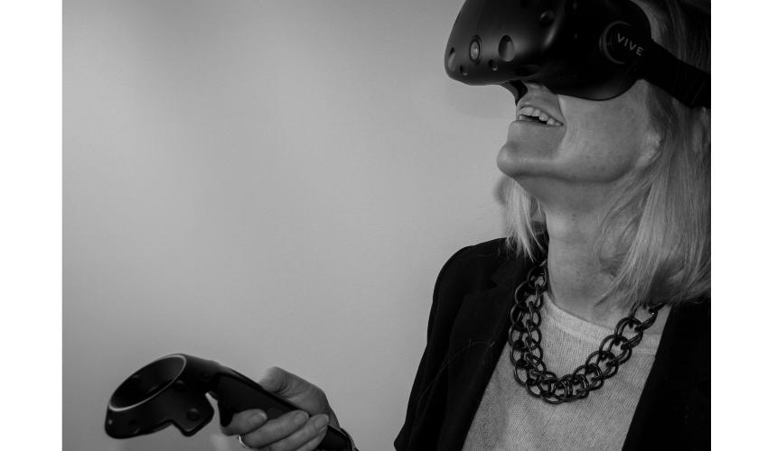 A black and white image of a light-haired woman smiling while wearing a virtual reality headset over her eyes, with her head tilted back and holding a controller