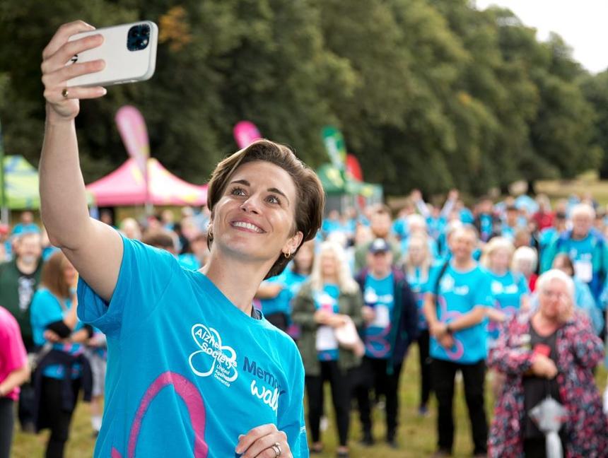 Vicky McClure wears a blue Memory Walk T-shirt and holds up her phone in front of a crowd of people to take a selfie