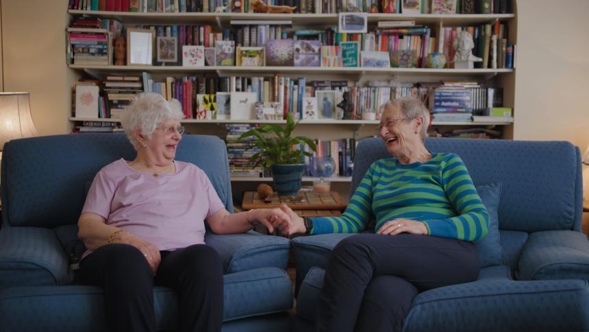 Hilary and Pauline sit in armchairs, holding hands and smiling