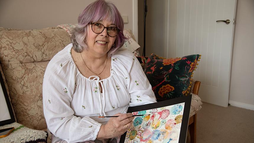 Gill Taylor sits on her sofa in a white blouse, she is holding a framed artwork she created