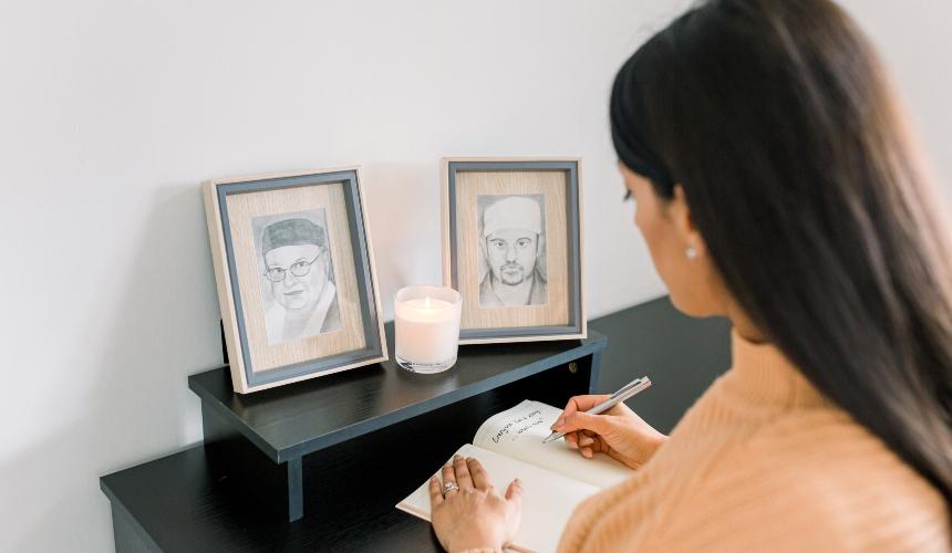 Karan Jutlla writing in a notebook, with a candle and some drawings in frames in front of her on a table