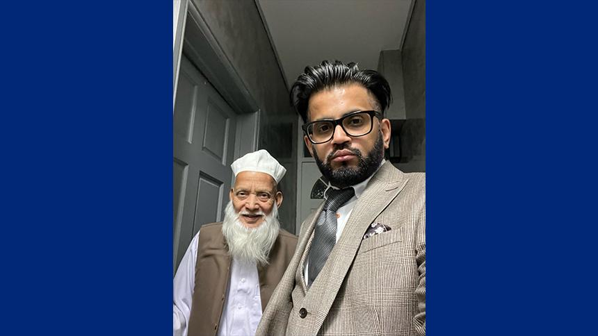 Muhammad stands next to his son Shahbaz in their home, they're both smiling at the camera which Shahbaz is holding to take a picture