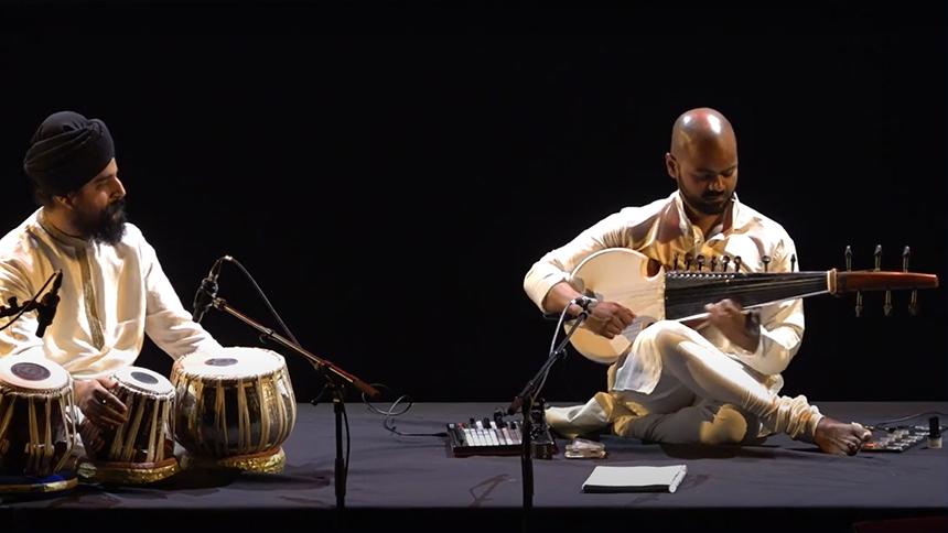A man plays a sarod (a stringed instrument used in North Indian classical music) on stage accompanied by a fellow musician on hand drums