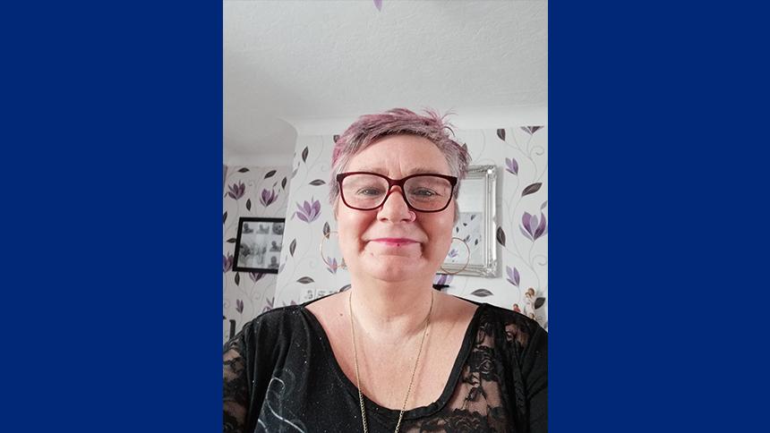 Michelle Nelson-Greensmith stands in her living room. She is looking at the camera wearing glasses and a black blouse.