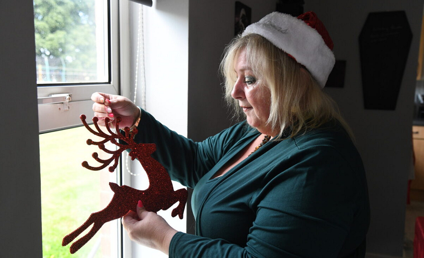 Anita standing by a window in a Christmas hat while holding a reindeer decoration