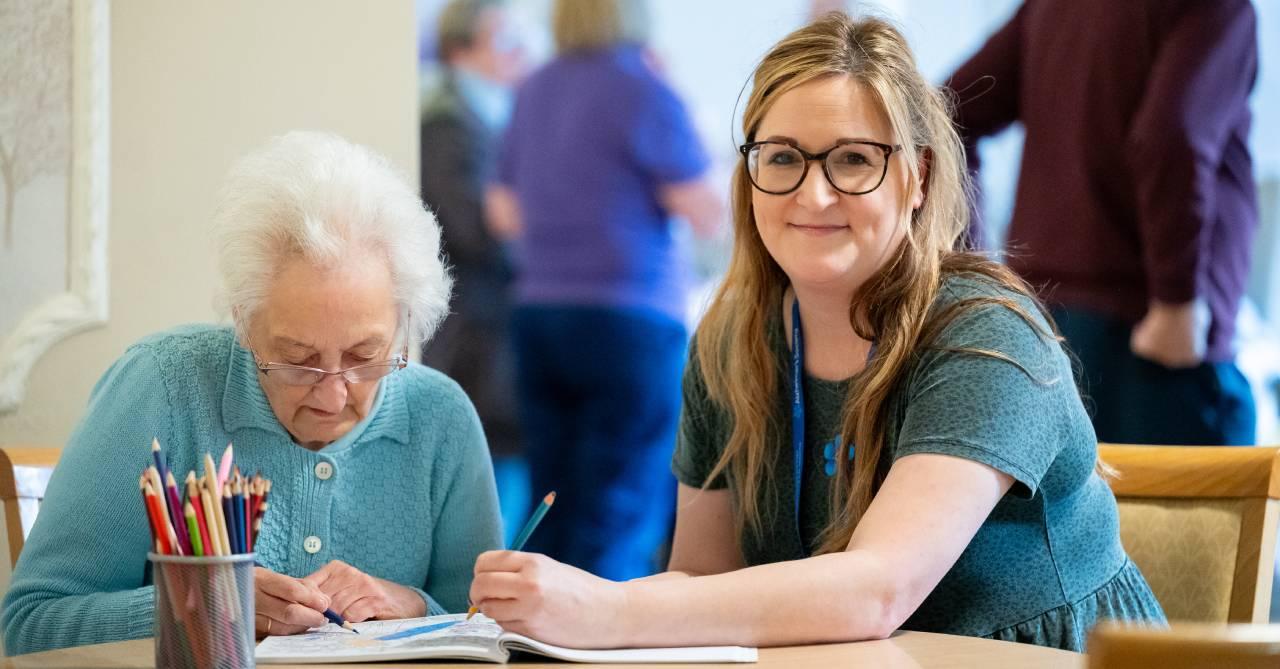 Researcher & care home resident living with dementia sat at a table in care home doing some colouring in