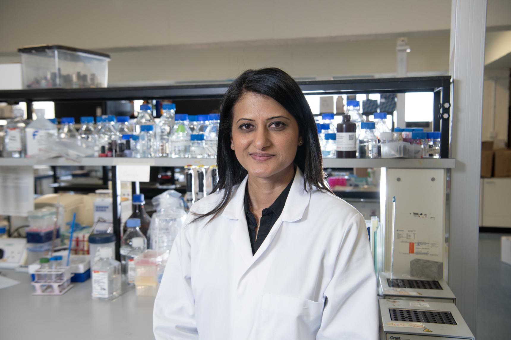 A scientist stands in the lab wearing a lab coat, smiling at the camera