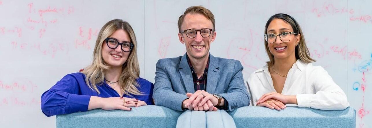Two women and a man stand together in front of a whiteboard.