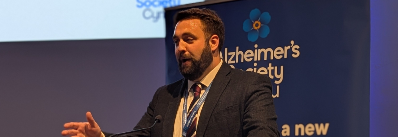 A man in a suit speaks on stage in front of an Alzheimer's Society banner.
