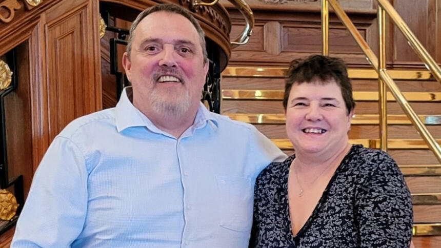 A man with short dark hair and a white beard wearing a light blue shirt and a woman with short brown hair wearing a black top with grey flowers