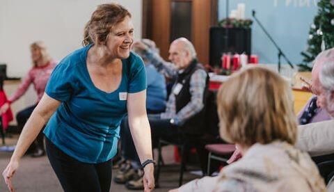 A woman in a sports-top moves across a room of seated people.