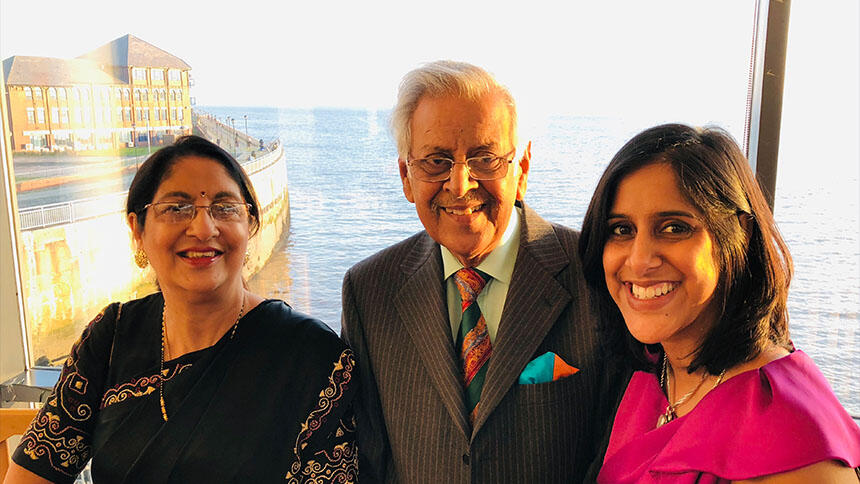Nupur is wearing a pink top, next to her is her dad Bulu who is wearing a suit and tie, and her mum Protima wearing a black top. They are stood next to a window, overlooking the sea and it's very sunny outside.