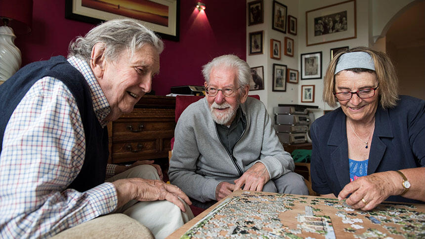 Three people, two men and a woman, huddled around a low table, doing a jigsaw.