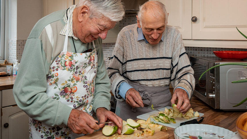 Two men stood in a kitchen, they're next to a table cutting up cooking apples.