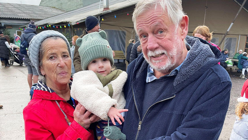 Ellen and her husband Malcolm. Ellen is wearing a red coat and grey hat. Malcolm is wearing a blue coat and holding a toddler.