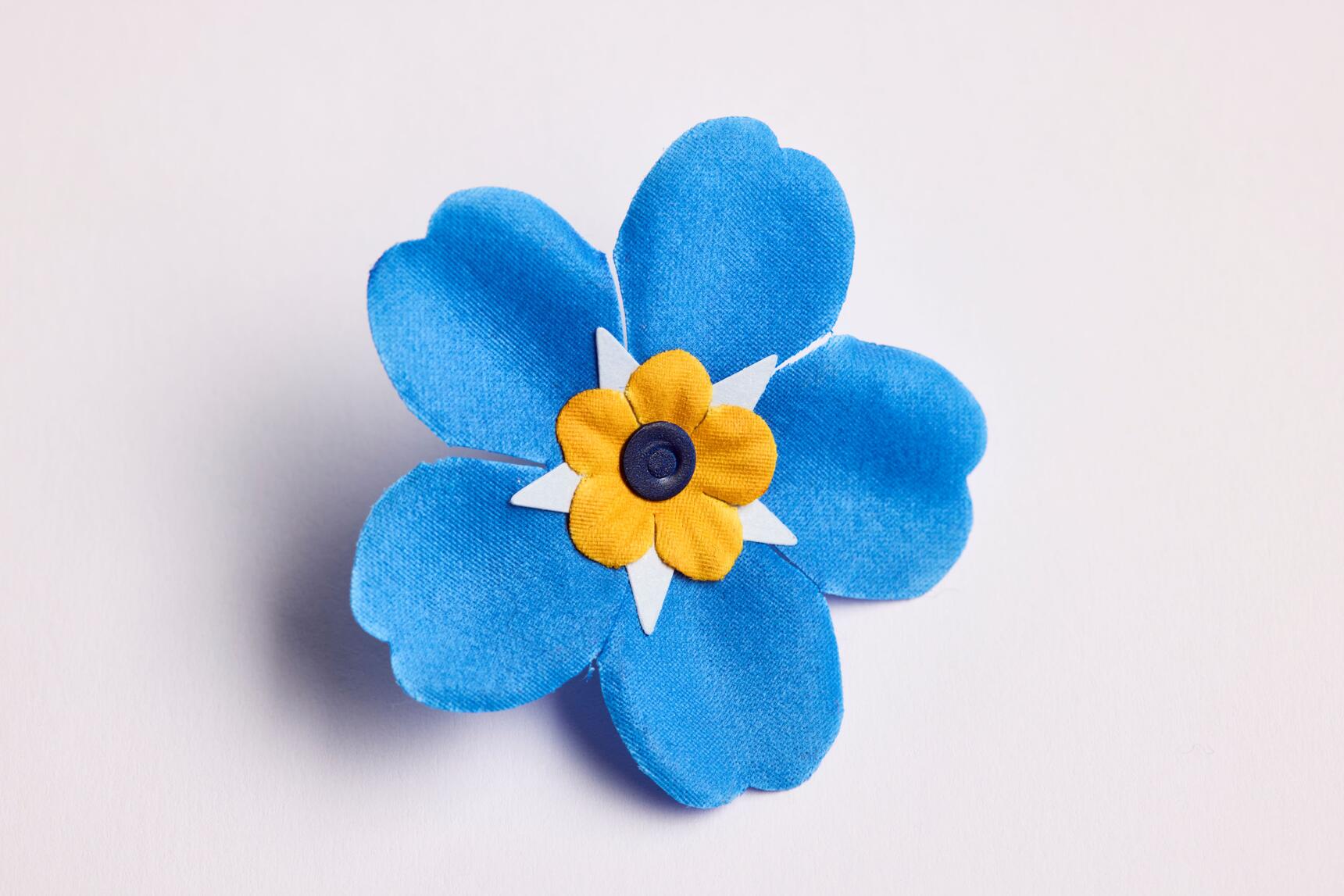 A fabric forget-me-not badge with blue petals, a yellow centre with a plastic dot