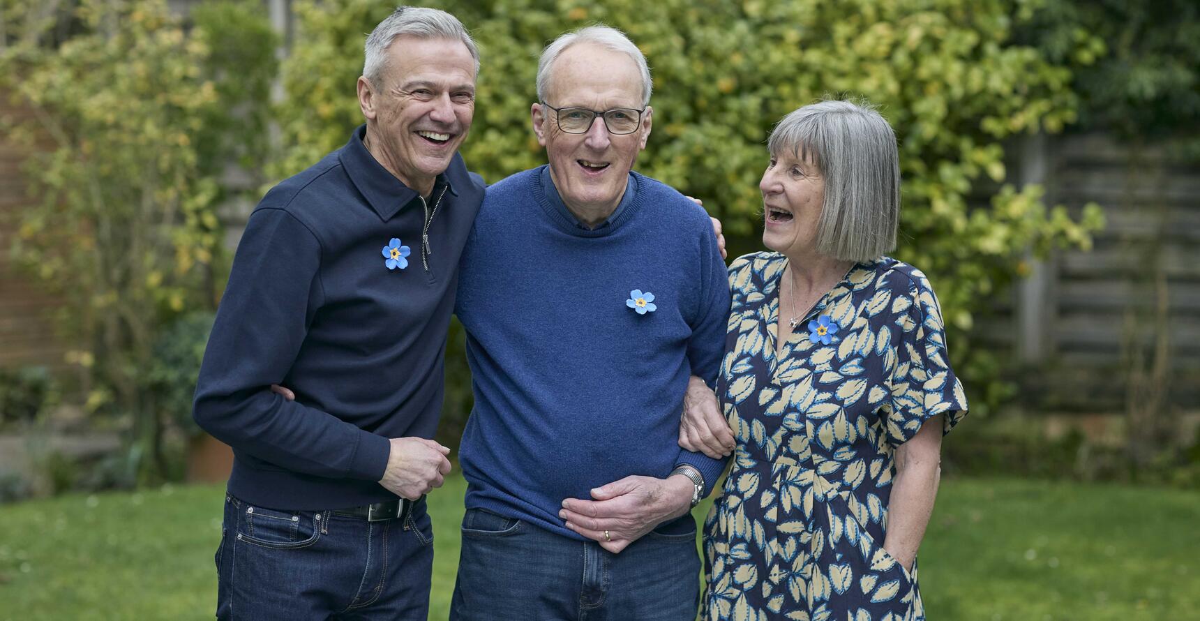 A man in blue shirt and jeans, a man with glasses, a blue jumper and jeans and a woman in a blue patterned dress in a garden. All have grey hair and are wearing forget me not badges