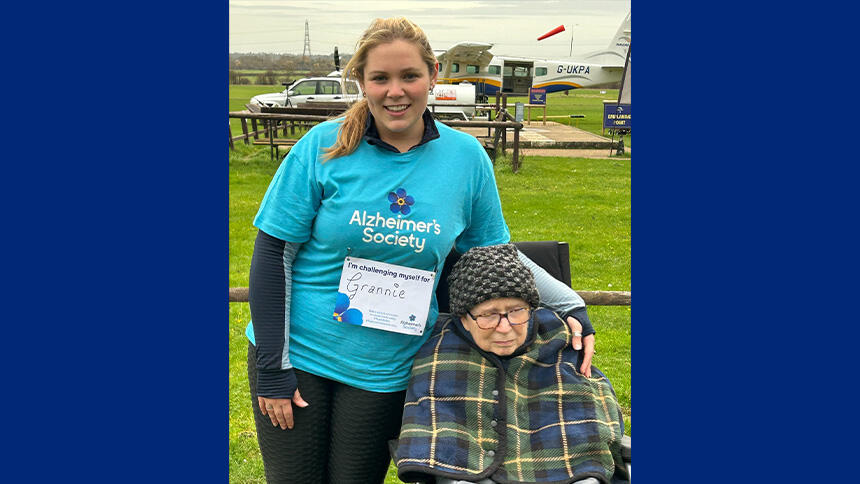 Abi-Lou is outside with her arm around her grannie Jean, there's an airfield behind them. She's wearing a blue Alzheimer's Society t-shirt and her grannie is wrapped in a tartan blanket.
