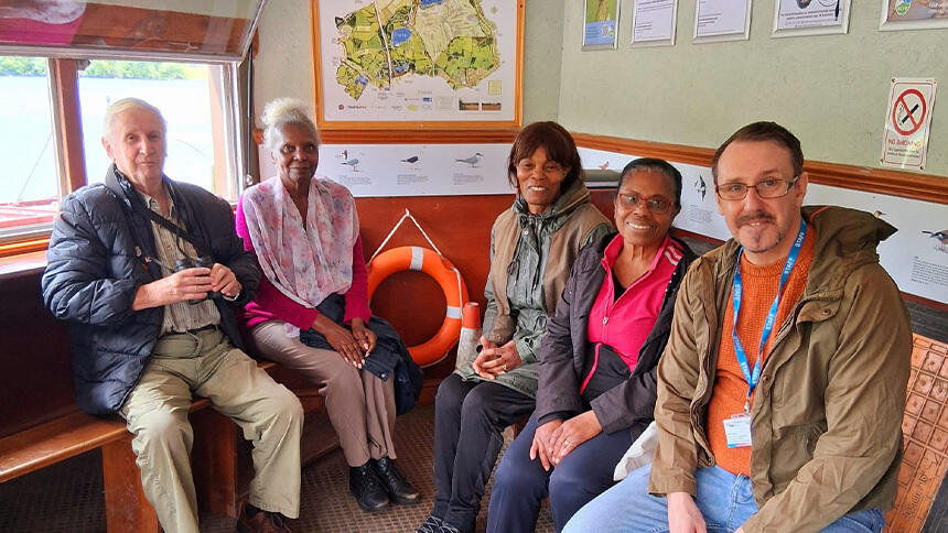 Five people sat on wooden benches, behind them are posters, a map and an orange lifebuoy