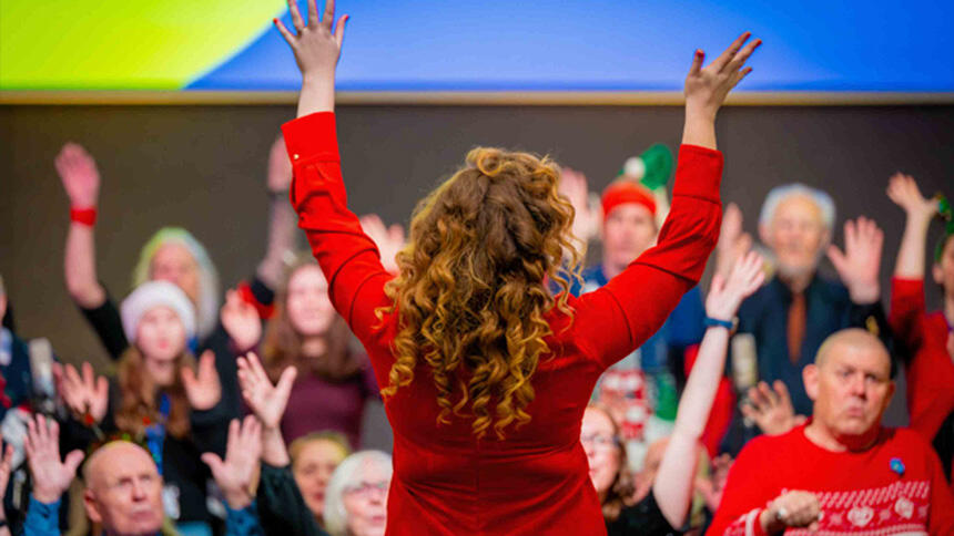 Choir of around 30 people holding their hands in the air, in front of them is a choir leader wearing red, with her hands in the air