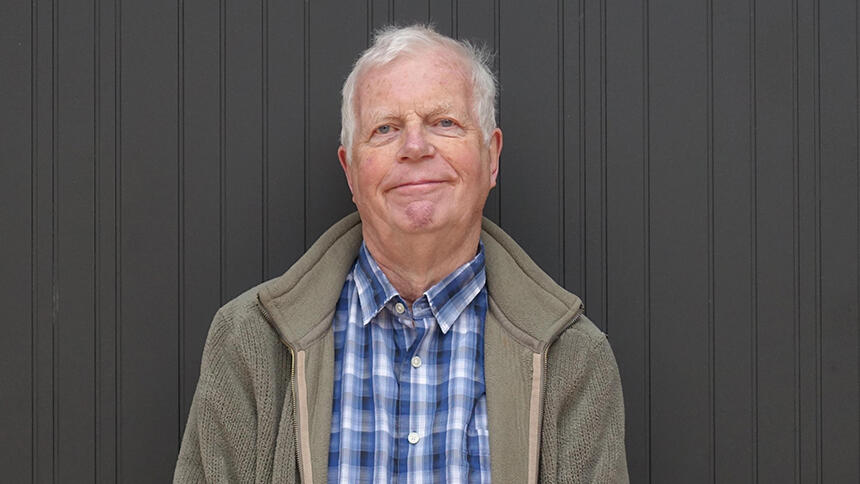 John has short white hair and he is wearing a blue checked shirt and a beige fleece jacket.