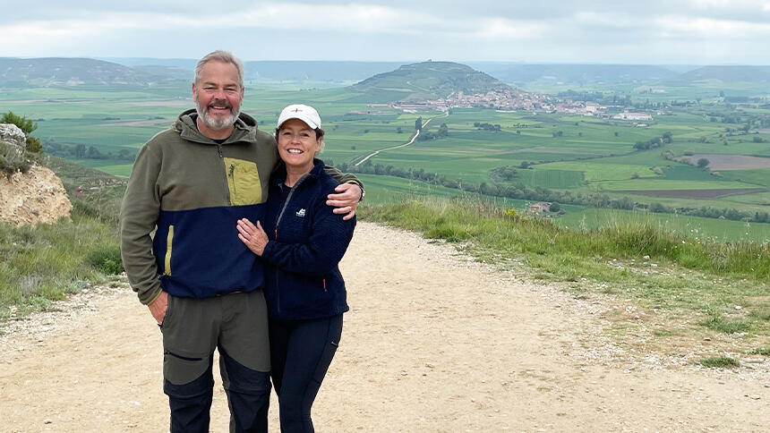 Kevin and Victoria are stood on a stony track of the Camino, with hilly scenery behind them