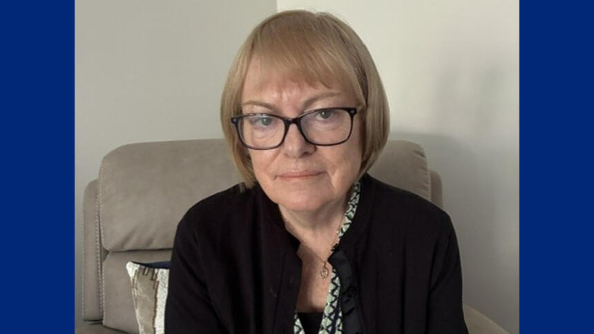 Karen has black-framed glasses and is wearing a black cardigan, she's sat on a beige armchair.