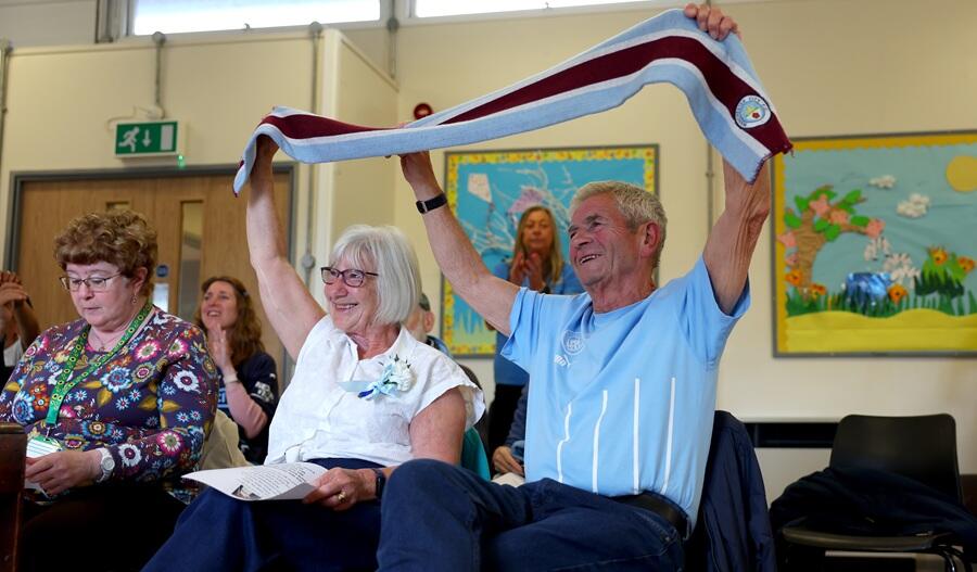 Two football supporters seated at a Dementia Cafe holding a football scarf above their heads