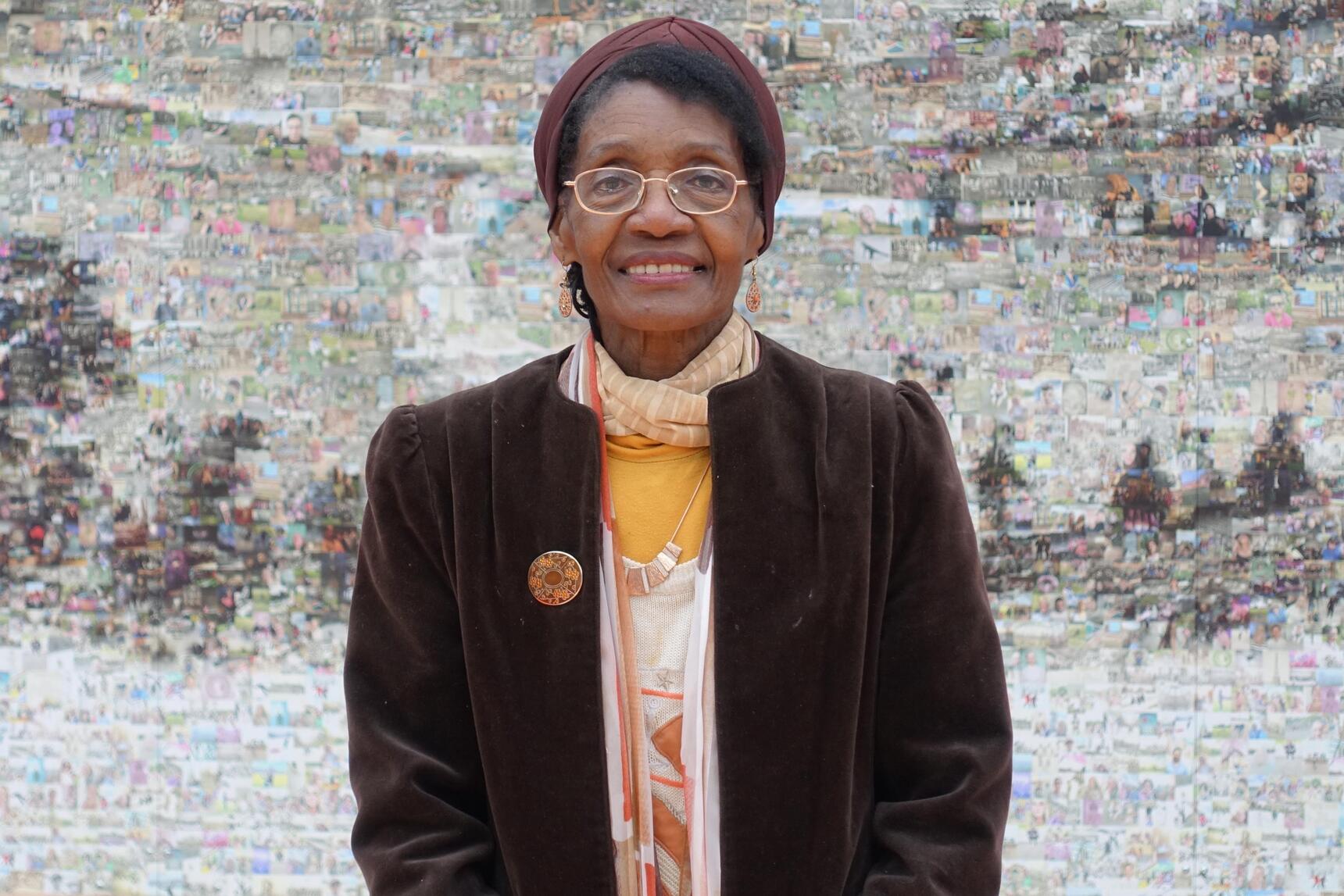 A woman wearing a brown jacket and glasses smiling in front of a piece of art